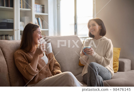 Happy european young women in casual with cups drink coffee, communication, sit on sofa in living room Happy european young women in casual with cups drink coffee, communication, sit on sofa in living room 98476145