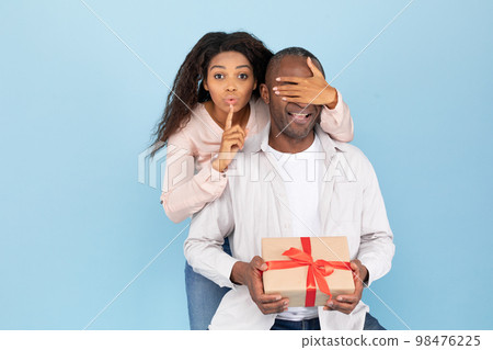 Happy anniversary. Portrait of young black woman covering her husband's eyes, making hush sigh, giving him gift box Happy anniversary. Portrait of young black woman covering her husband's eyes, making hush sigh, giving him gift box 98476225