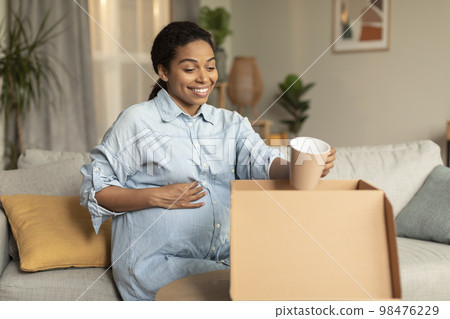 Excited pregnant black woman unpacking cardboard moving box, holding cup and smiling, sitting on couch at home Excited pregnant black woman unpacking cardboard moving box, holding cup and smiling, sitting on couch at home 98476229