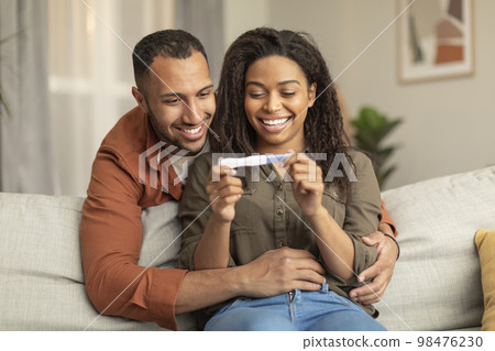 Happy african american couple holding positive pregnancy test and smiling, man hugging woman on sofa at home Happy african american couple holding positive pregnancy test and smiling, man hugging woman on sofa at home 98476230
