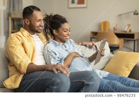 Excited african american pregnant couple using digital tablet, choosing and purchasing clothes for baby in internet Excited african american pregnant couple using digital tablet, choosing and purchasing clothes for baby in internet 98476235