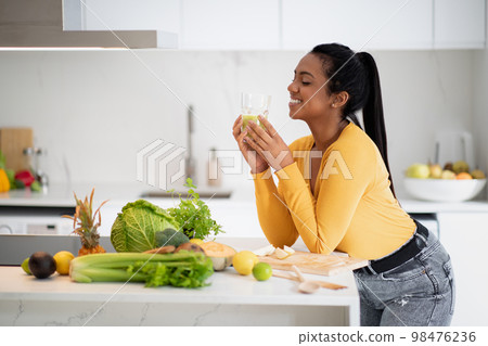 Smiling young african american female drinking glass of smoothie at table with organic vegetables, enjoy diet Smiling young african american female drinking glass of smoothie at table with organic vegetables, enjoy diet 98476236