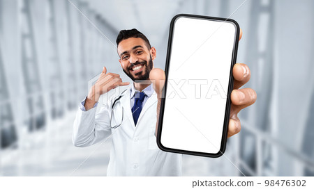 Middle eastern male doctor showing blank smartphone and making call me gesture Middle eastern male doctor showing blank smartphone and making call me gesture 98476302