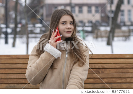 Beautiful girl in sheepskin coat sits on bench in winter park and talks on smartphone. Serious young woman with gadget. 98477631