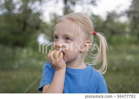 Little blond girl in blue t-shirt bite green apple . Child walks in park or garden. Close-up portrait of little girl. Little blond girl in blue t-shirt bite green apple . Child walks in park or garden. Close-up portrait of little girl. 98477791