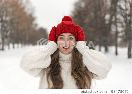 Beautiful young girl in wool red hat and mittens smiles straight into camera. Woman in knitted clothes in winter forest Beautiful young girl in wool red hat and mittens smiles straight into camera. Woman in knitted clothes in winter forest 98478071