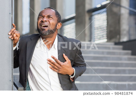 Heart attack, African American man has severe chest pain, businessman outside office building on stairs writhing in chest pain. 98481874
