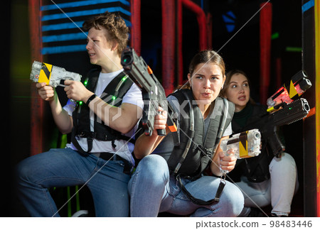 Two girls and one guy sitting and hiding behid cover while playing lasertag Two girls and one guy sitting and hiding behid cover while playing lasertag 98483446