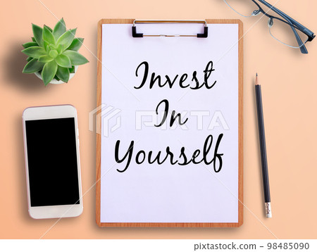 Top view text invest in yourself on clipboard,smartphone,pot plant and pencil on orange background. 98485090