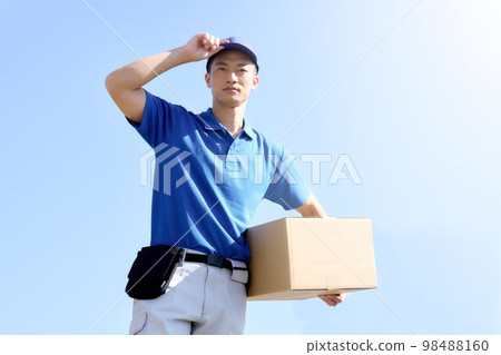 A delivery man holding a package under the blue sky and putting his hand on his hat Transportation Logistics Business image 98488160