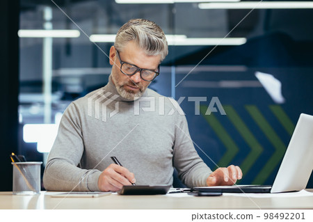 Serious thinking businessman inside office at work with laptop, senior gray haired man working sitting at desk. 98492201