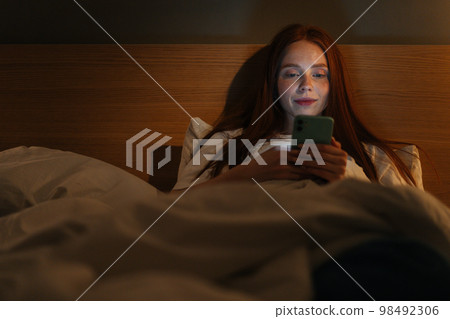 Portrait of happy smiling young woman using smartphone, looking on screen, typing online message on social media, lying on bed late at night, bedside lamp lighting with warm yellow light. Portrait of happy smiling young woman using smartphone, looking on screen, typing online message on social media, lying on bed late at night, bedside lamp lighting with warm yellow light. 98492306