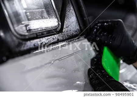 Employee of a car detailing studio applies a colorless protective film covering the paintwork of a white car Employee of a car detailing studio applies a colorless protective film covering the paintwork of a white car 98495901