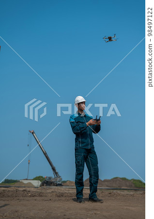 A man in a helmet and overalls controls a drone at a construction site. The builder carries out technical oversight. A man in a helmet and overalls controls a drone at a construction site. The builder carries out technical oversight. 98497122