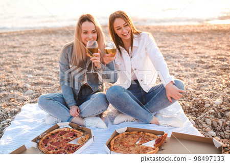 Two young beautiful girlfriends having fun with pizza and wine at beach over sea shore outdoors in sun light. Summer vacation season. Friendship. Happiness. Sisters spend time together at coast line. 98497133