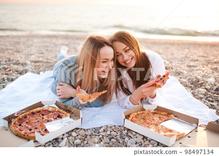 Two young beautiful girlfriends having fun with pizza and wine at beach over sea shore outdoors in sun light. Summer vacation season. Friendship. Happiness. Sisters spend time together at coast line. Two young beautiful girlfriends having fun with pizza and wine at beach over sea shore outdoors in sun light. Summer vacation season. Friendship. Happiness. Sisters spend time together at coast line. 98497134