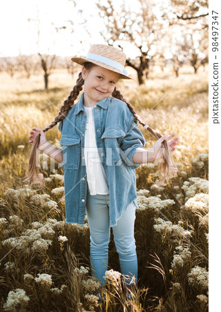 Happy kid girl 4-5 year old wearing straw hat and decnim jacket with pants posing in flower meadow outdoors. Looking at camera. Spring season. Childhood. Happiness. 98497347