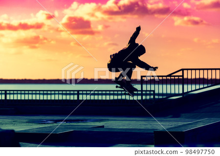 Unrecognizable teenage boy silhouette showing high jump tricks on scooter against 98497750
