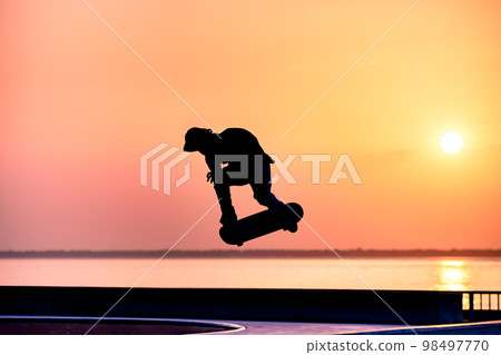 Unrecognizable teenage boy silhouette showing high jump tricks on scooter against 98497770