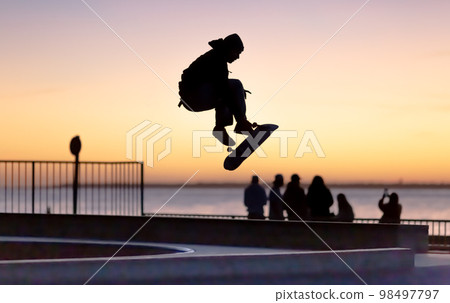 Unrecognizable teenage boy silhouette showing high jump tricks on scooter against 98497797