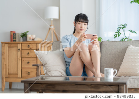 坐在客廳沙發上用智能手機看視頻的女高中生 坐在客廳沙發上用智能手機看視頻的女高中生 98499833