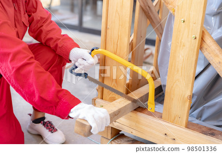 A worker is sawing wood to assemble a crate for moving an industrial machine. A woman in red mechanic coveralls hand holding a bow saw cutting the wooden plank of a crate. Safety in workplace concept. A worker is sawing wood to assemble a crate for moving an industrial machine. A woman in red mechanic coveralls hand holding a bow saw cutting the wooden plank of a crate. Safety in workplace concept. 98501336