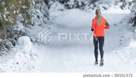 Running in winter, rear view of a female runner on a snowy path in a park or forest 98501372