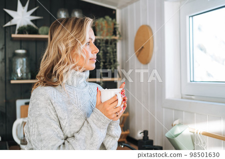 Young woman in grey sweater with cup of hot drink in hands looking at window on kitchen at house 98501630