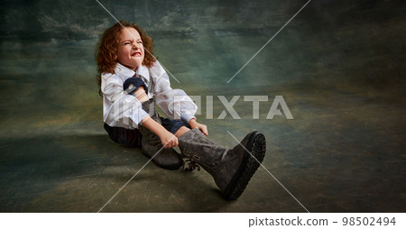 Wide angle view of little cute kid with curly hair sitting on floor over dark green background. Concept of childhood, comparison of eras, retro, vintage, emotions Wide angle view of little cute kid with curly hair sitting on floor over dark green background. Concept of childhood, comparison of eras, retro, vintage, emotions 98502494