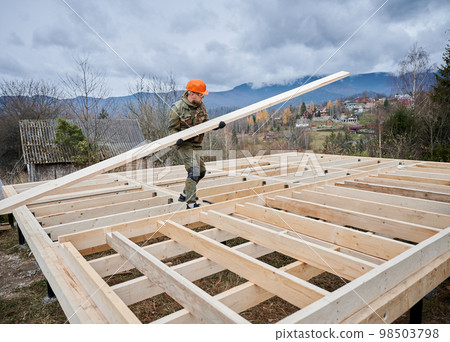 Man worker building wooden frame house on pile foundation. Carpenter carrying wooden board. Carpentry concept. 98503798