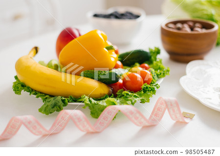 Measuring tape on the background of vegetables and fruits. The concept of healthy food and weight loss Measuring tape on the background of vegetables and fruits. The concept of healthy food and weight loss 98505487