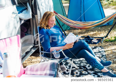 Adult woman relaxing, sitting in the travel chair and reading book on the background of camper car vehicle. Female living on camper car and travel the world. Caravan car Vacation. Adventure Is Ageless Adult woman relaxing, sitting in the travel chair and reading book on the background of camper car vehicle. Female living on camper car and travel the world. Caravan car Vacation. Adventure Is Ageless 98505561
