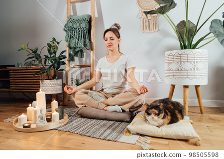 Young healthy woman practicing meditation at home, sitting in lotus position on floor cushion with relaxed cat pet in modern Scandi interior home. Meditating, relaxed, eyes closed. Selective focus 98505598