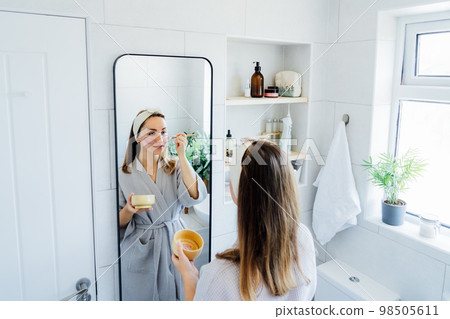 Young woman in bathrobe looking in the mirror and applying natural cosmetic clay mask on her face in bathroom. Cosmetic procedures for teens skin care at home. Beauty self-care. Selective focus. 98505611