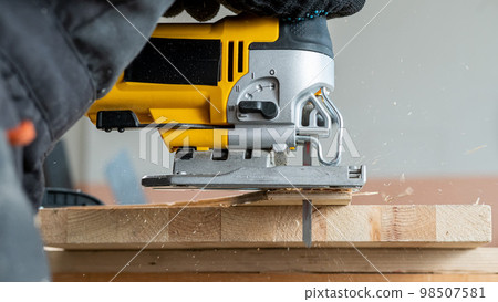 Close-up of a man cutting a wooden plank with an electric jigsaw in a workshop. Close-up of a man cutting a wooden plank with an electric jigsaw in a workshop. 98507581