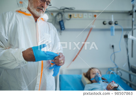 Doctor in protective coverall putting off surgical gloves after examining little girl. Doctor in protective coverall putting off surgical gloves after examining little girl. 98507863