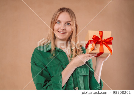 Happy young blonde woman 20s wears green denim jacket hold shake red present box with gift ribbon bow try to guess what inside isolated on plain pastel light beige background studio portrait 98508201