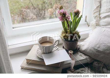 Feminine spring composition. Cup of coffee, books and blank greeting card mockup near window. Vintage Easter styled photo. Moody floral composition. Potted pink hyacinth flowers. Linen cushion. 98509404
