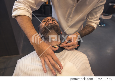Barber with tattoed arm carefully shaving the client 98509679