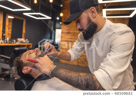 Barber with tattoed arm carefully shaving the client 98509681