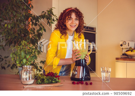 Curly-haired girl looking involved while making smoothie in the kitchen Curly-haired girl looking involved while making smoothie in the kitchen 98509692