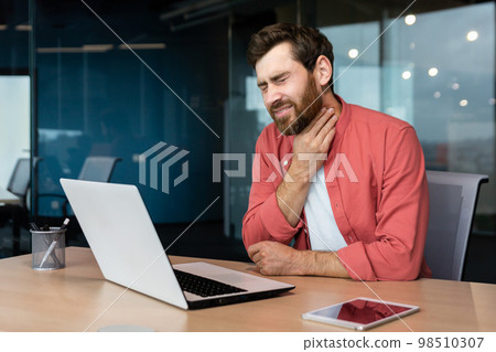 A man with a sore throat works inside the office, a businessman at work has a sore throat flu and cold, uses a laptop at work. A man with a sore throat works inside the office, a businessman at work has a sore throat flu and cold, uses a laptop at work. 98510307