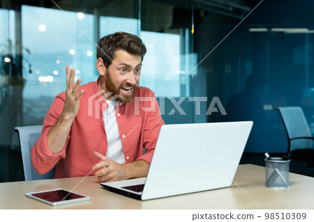 Angry and nervous businessman talking on video call remotely, man shouting to colleagues online, boss in shirt working inside modern office using laptop at work. Angry and nervous businessman talking on video call remotely, man shouting to colleagues online, boss in shirt working inside modern office using laptop at work. 98510309