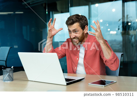 Angry and nervous businessman talking on video call remotely, man shouting to colleagues online, boss in shirt working inside modern office using laptop at work. Angry and nervous businessman talking on video call remotely, man shouting to colleagues online, boss in shirt working inside modern office using laptop at work. 98510311