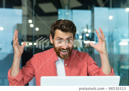Angry and nervous businessman talking on video call remotely, man shouting to colleagues online, boss in shirt working inside modern office using laptop at work. Angry and nervous businessman talking on video call remotely, man shouting to colleagues online, boss in shirt working inside modern office using laptop at work. 98510313