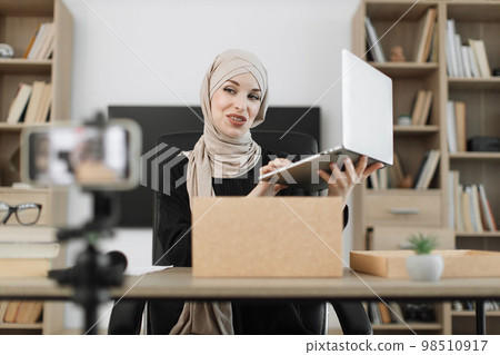 Muslim woman recording video on phone camera while unpacking box with new wireless laptop. Muslim woman recording video on phone camera while unpacking box with new wireless laptop. 98510917