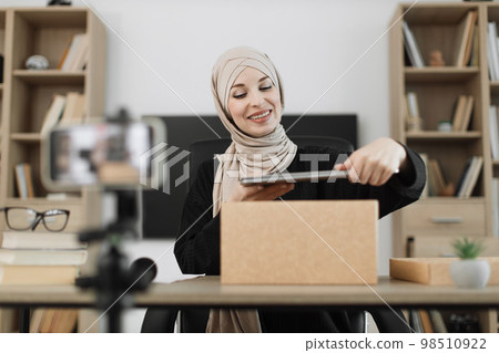 Muslim woman recording video on phone camera while unpacking box with new wireless laptop. Muslim woman recording video on phone camera while unpacking box with new wireless laptop. 98510922