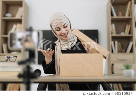 Excited smiling muslim woman in casual wear and hijab recording video on camera while unpacking gift 98510924