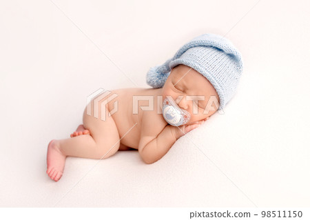 A cute newborn boy in the first days of life sleeps naked with a white pacifier in his mouth on white fabrics in blue knitted woolen cap. Studio professional macro photography, portrait of a newborn. 98511150