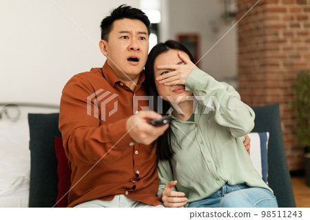 Scared asian woman and man with remote control switching channel on tv, watching horror film in living room interior Scared asian woman and man with remote control switching channel on tv, watching horror film in living room interior 98511243